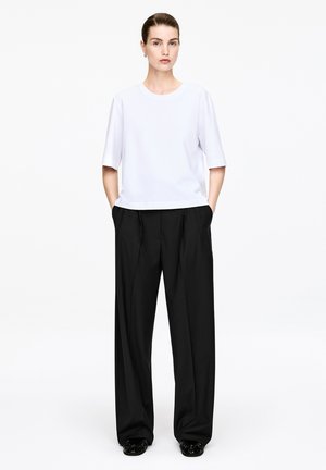 Kvinde stående med hænderne i lommerne iført en hvid kortærmet top, brede sorte bukser og sorte sko mod en ensfarvet baggrund.