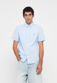 Ung mann iført en lys blå kortermet skjorte med knapper og lys blå jeans, stående med den ene hånden i lommen mot en hvit bakgrunn.