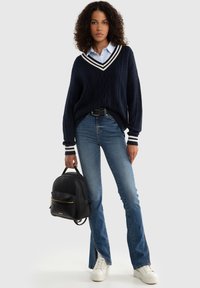 Maglione navy a trama cable knit con strisce bianche a V, camicia azzurra sotto, jeans azzurri chiari, sneakers bianche e uno zaino nero.