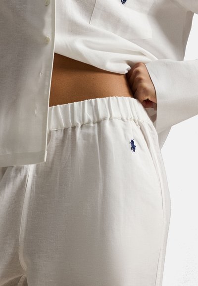 Witte broek met elastische taille en een klein geborduurd logo op de heup, gecombineerd met een bijpassend wit hemd met knopen, waarbij de buik zichtbaar is.