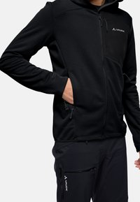 Giacca di pile nera con zip frontale, tasche laterali e un pannello in tessuto nero a contrasto. Presenta un logo sul petto e sul cappuccio.