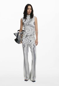 Wijd uitlopende jumpsuit in het wit met zwarte draaiende patronen, met een hoge hals en een getailleerde corsage. Model houdt een grote grijze tas met een print vast.