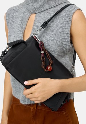 Osoba ubrana w szary bezrękawnik dzianinowy i brązowe spodnie trzyma czarną torebkę z ozdobną liną i dołączonym bursztynowym koralikiem.