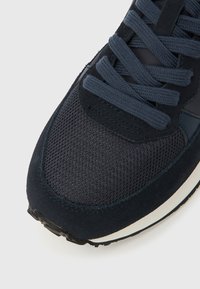 Tumesinine toss, mille varbaküljel on võrgukangas, pealdised seemisnahast, paksud paelad ning valge keskmine tald ja must põhjaosa, valgel taustal nurga all.