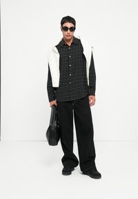 Czarna koszula w kratkę, przesadnie duże czarne spodnie, biała bluza zarzucona na ramiona, trzymająca czarną torbę, nosząca okulary przeciwsłoneczne, stojąca na tle gładkiej ściany.