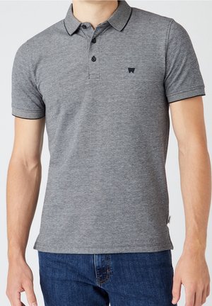 Mann trägt ein eng anliegendes graues Polohemd mit kurzen Ärmeln, zwei Knöpfen und einem dezenten schwarzen Logo auf der Brust, kombiniert mit blauen Jeans.