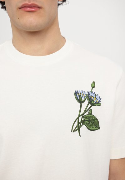 Cremfarbendes Baumwoll-T-Shirt mit einem grünen und blauen Blumenstickdesign im linken Brustbereich. Rundhalsausschnitt, kurze Ärmel.