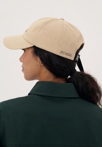 Beżowa czapka baseballowa wykonana z bawełnianego tkaniny typu twill, z regulowanym paskiem i akcentem tekstowym z tyłu. Nosi ją osoba w ciemnozielonej koszuli z kołnierzykiem.