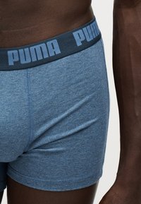 Κοντινό πλάνο ενός ατόμου που φορά μπλε boxer briefs της Puma με σκούρα μπλε ζώνη μέσης που εμφανίζει το λογότυπο της Puma.