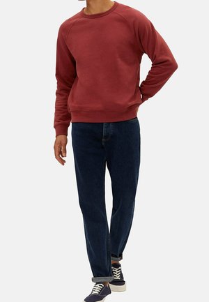 Persona che indossa una felpa rossa a maniche lunghe, jeans blu scuro e sneakers nere con lacci bianchi, in piedi contro uno sfondo semplice.