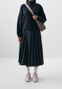 Černá fleecová mikina na zip, plisovaná tmavá sukně, růžové a šedé tenisky, bílé ponožky s černým logem a hnědá crossbody taška.