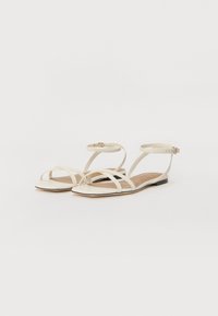 Cream flat sandals with multiple crossed straps and ankle buckle. Smooth material, square toe design, and light brown insoles.