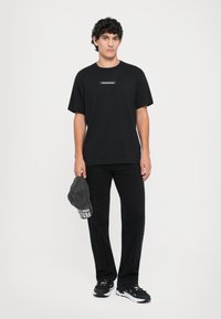 Svart bomullst-skjorte med korte ermer og et sentralt logo-patch, kombinert med svarte jeans og en grå caps. Svarte joggesko fullfører antrekket.