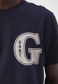 Camiseta de algodón azul marino con una gran letra blanca "G" y la palabra "GANT" al lado en un estilo de fuente moderno. Mangas cortas, corte clásico.