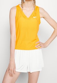 Oranžový bezrukávový sportovní top s véčkovým výstřihem a texturovanou tkaninou, kombinovaný s bílými šortkami; na hrudi je logo Nike.