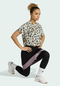 T-shirt con stampa leopardata, tagliata, in tonalità beige e nero, abbinata a leggings neri. Una fascia di resistenza rosa è posizionata attorno alle cosce. Scarpe da ginnastica bianche indossate.