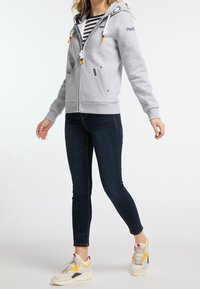 Felpa grigia chiara con zip e cappuccio, dotata di lacci a contrasto, tasche laterali e polsini a costine, indossata sopra una maglietta a righe e jeans scuri.