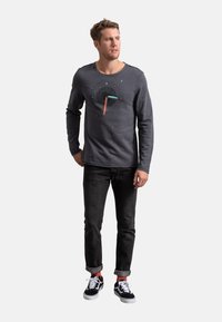 Maglietta a maniche lunghe grigio scuro con un design grafico circolare in nero, turchese e corallo, abbinata a jeans neri e scarpe da ginnastica nere.
