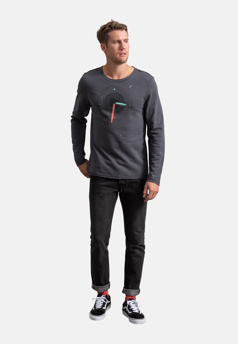 Maglietta a maniche lunghe grigio scuro con un design grafico circolare in nero, turchese e corallo, abbinata a jeans neri e scarpe da ginnastica nere.