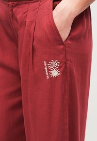 Χέρι ατόμου στην τσέπη μαρόν παντελονιού με διπλή κεντητή απεικόνιση ήλιου και λογότυπο Quiksilver στην πλευρική ραφή.