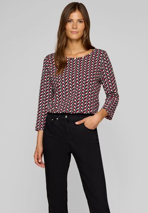 Mehrfarbige Langarmbluse mit geometrischem Muster in Rot, Weiß und Schwarz; kombiniert mit schwarzen Hosen und einem lässigen Schnitt.