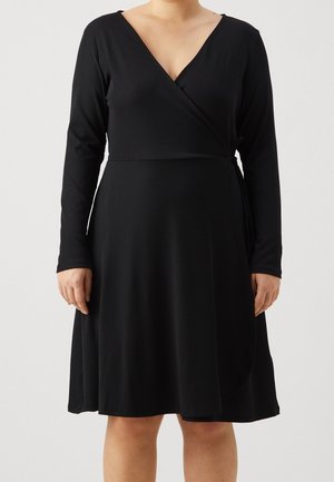 Femme portant une robe portefeuille noire à manches longues, longueur genou, avec un décolleté en V, sur un fond clair uni.