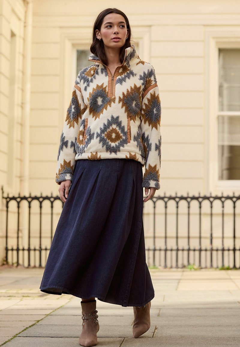 Fleece-Pullover mit geometrischem Muster in Creme, Braun und Grau; verfügt über einen halbhohen Reißverschluss am Kragen und gerippte Bündchen. Kombiniert mit einem langen Jeansrock.