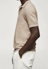 Polo à manches courtes en tissu beige clair avec une finition texturée, arborant un col classique et un design ajusté, associé à un pantalon blanc.