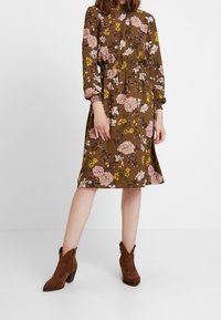 Braunes geblümtes Kleid mit langen Ärmeln, geraffter Taille und Manschetten. Rosa und gelbe Blumen auf einem gedämpften Hintergrund. Braune Knöchelstiefel.
