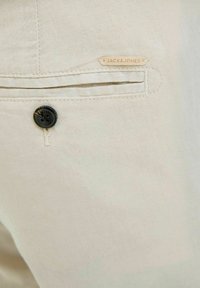 Jasnobeżowe spodnie z tylną kieszenią z czarnym guzikiem oraz małą skórzaną metką z logo "JACK&JONES". Gładka tekstura materiału.