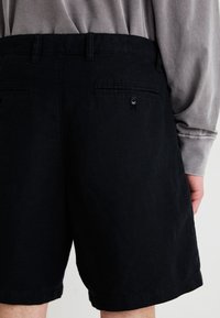 Schwarze Shorts aus einem strukturierten Stoff, mit einer Gesäßtasche mit Knopf. Das Design umfasst Gürtelschlaufen und einen geraden Saum.