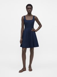 Femme aux cheveux courts portant une robe bleu marine sans manches, longueur genou, et des sandales plates marron, debout avec une main sur la hanche.