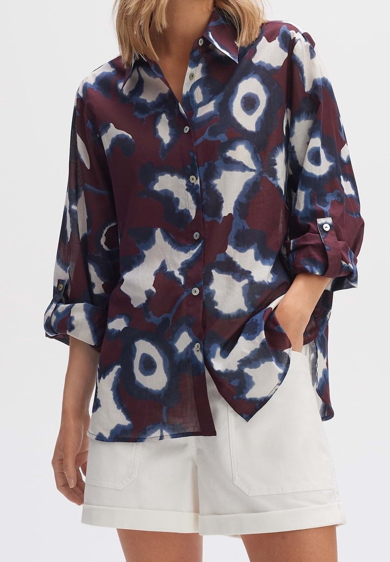 Femme portant une chemise ample à manches longues boutonnée avec des motifs abstraits bleus et blancs, et un short blanc retroussé, une main dans la poche.