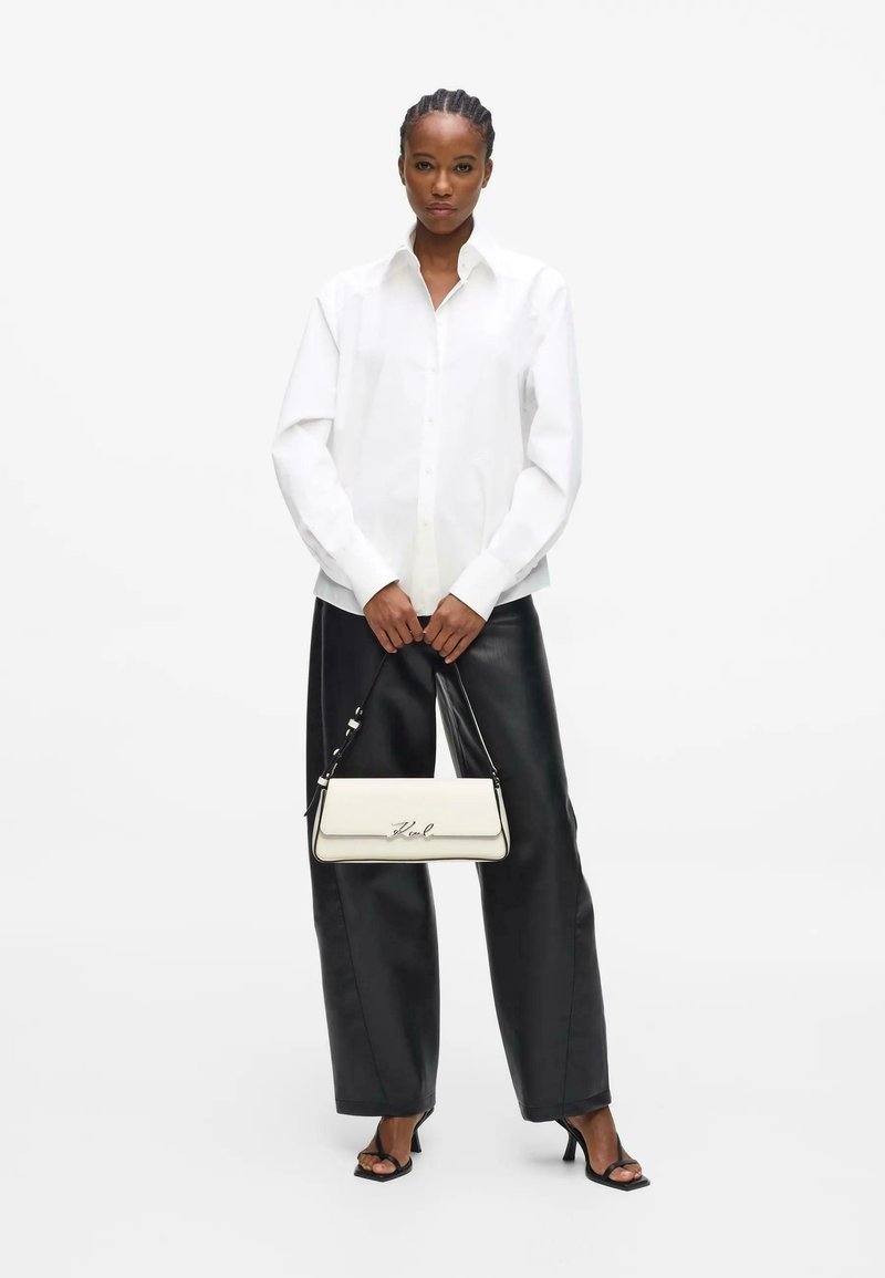 Femme portant une chemise blanche boutonnée et un pantalon noir à jambes larges, tenant un sac à main blanc, debout avec des pieds dans des sandales à talons noires.