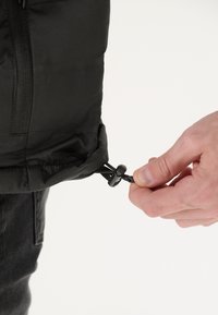 Giubbotto a sacco nero con finitura testurizzata. Una mano regolano il cordino all'orlo, dotato di un gancio di plastica per il serraggio.