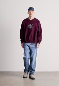 Bordový sweatshirt s šedým logem, doplněný světle modrými džíny a šedou obuví. Materiál je měkký a má volný střih.