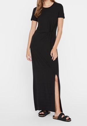 Zwarte maxi-jurk met korte mouwen, een zijsplit, een geraffineerd knoopdetail in de taille, gemaakt van een zachte, flexibele stof. Gecombineerd met zwarte sandalen.