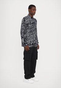 Schwarz-weißer gemusterter Langarmshirt mit verschiedenen Textdesigns, kombiniert mit schwarzen Cargo-Hosen und weißen Sneakers.