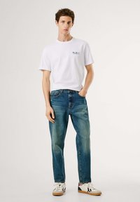 Hvid bomulds t-shirt med et lille blåt logo, kombineret med blå denim jeans. Modellen har hvid- og marineblå sneakers med beige såler på.