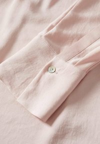 Tessuto della blusa rosa chiaro con una texture liscia, caratterizzato da una chiusura a bottone sul polsino e delicate pieghe lungo la manica.