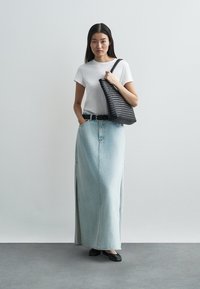 Gonna lunga in denim azzurro chiaro con spacchi alte fino alla coscia, abbinata a una t-shirt bianca e una borsa nera con trama. Scarpe basse nere completano il look.