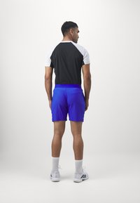 Sportoutfit met een zwart T-shirt met korte mouwen en blauwe textuurshorts, gecombineerd met witte sokken en schoenen, gezien vanuit de achterkant.