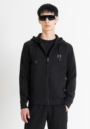 Sudadera negra con cremallera y capucha, que presenta un cierre frontal con cremallera, dos bolsillos laterales y un pequeño parche con logo en el pecho. Llevada con pantalones a juego.