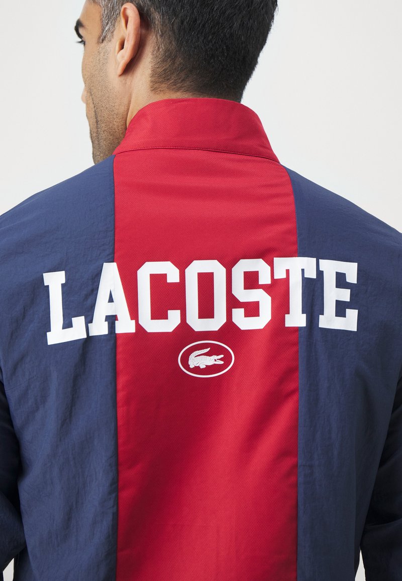 Veste colorée en rouge et marine avec un col haut, arborant un grand texte "LACOSTE" en blanc et un logo de crocodile au dos. Matériau léger.