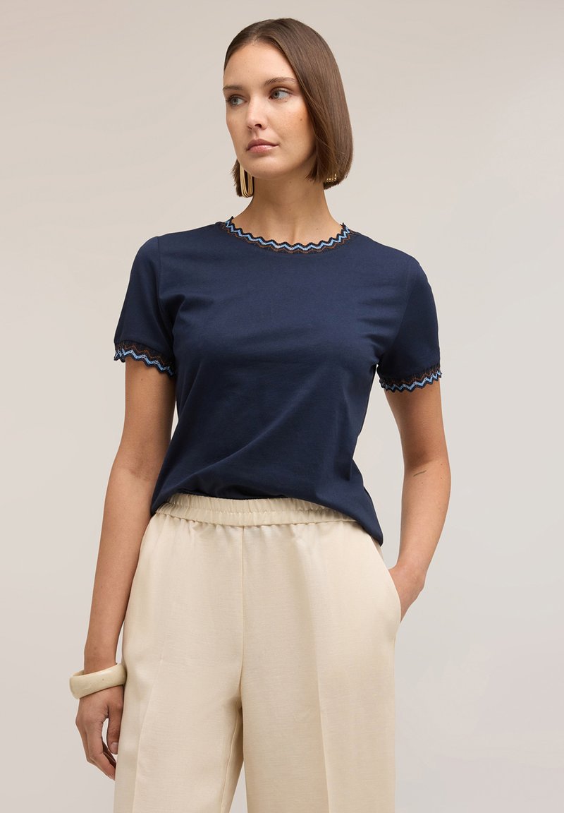 Mujer con cabello corto y castaño, vestida con una camiseta azul marino de manga corta con bordes ondulados y pantalones beige de talle alto, con una mano en el bolsillo.