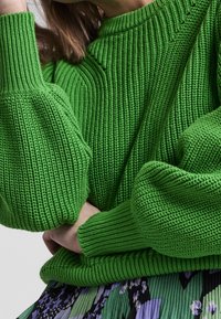 Pull vert en maille avec un motif texturé côtelé et des manches surdimensionnées. Une blouse à motif dépasse en dessous, ajoutant un contraste de couleurs.