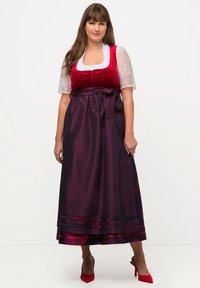 Perinteinen dirndl-mekko, jossa on punainen samettirintamerkki, valkoiset pitsihihat ja pitkä, tummanvioletti hame, jossa on kukkakoristeita ja vyötärökaari.