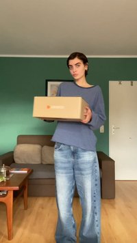 Caja de cartón sostenida en las manos, con un logo, frente a una persona vestida con un suéter azul a rayas y jeans de pierna ancha de lavado claro.