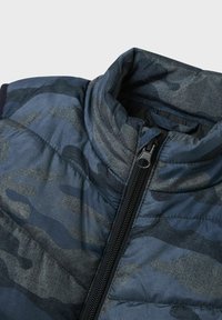 Puffer jakna s uzorkom kamuflaže u tamnoplavoj i sivoj boji, s visokim ovratnikom i crnim patentnim zatvaračem. Ima teksturiranu tkaninu i quilted dizajn.