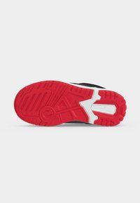 Semelle d'une chaussure de sport noire et rouge avec des rainures profondes et des détails blancs sur un fond gris clair uni.